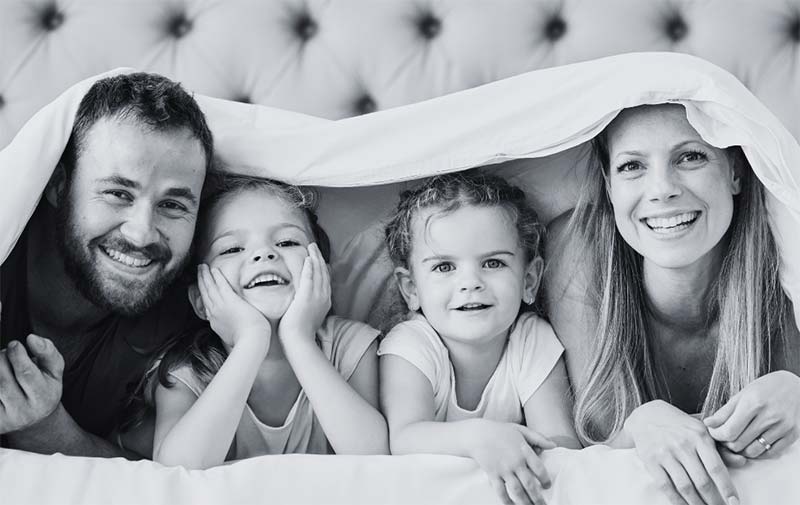 Eine Familie mit zwei Kindern liegt unter einer Bettdecke und freut sich